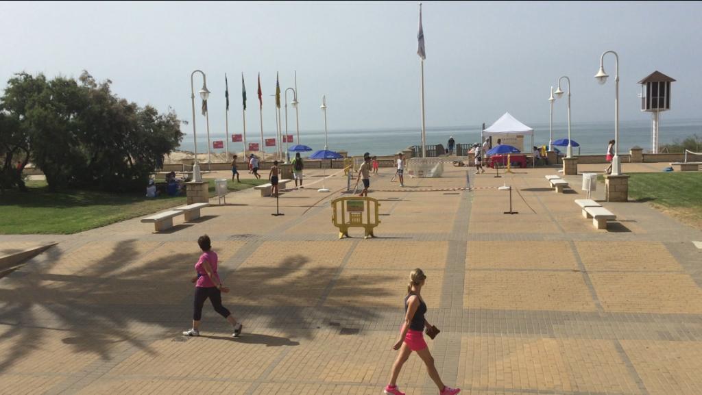 Vista del paseo marítimo de Islantilla en un día soleado, con personas caminando, banderas ondeando y el mar de fondo, creando un ambiente relajante junto a la playa