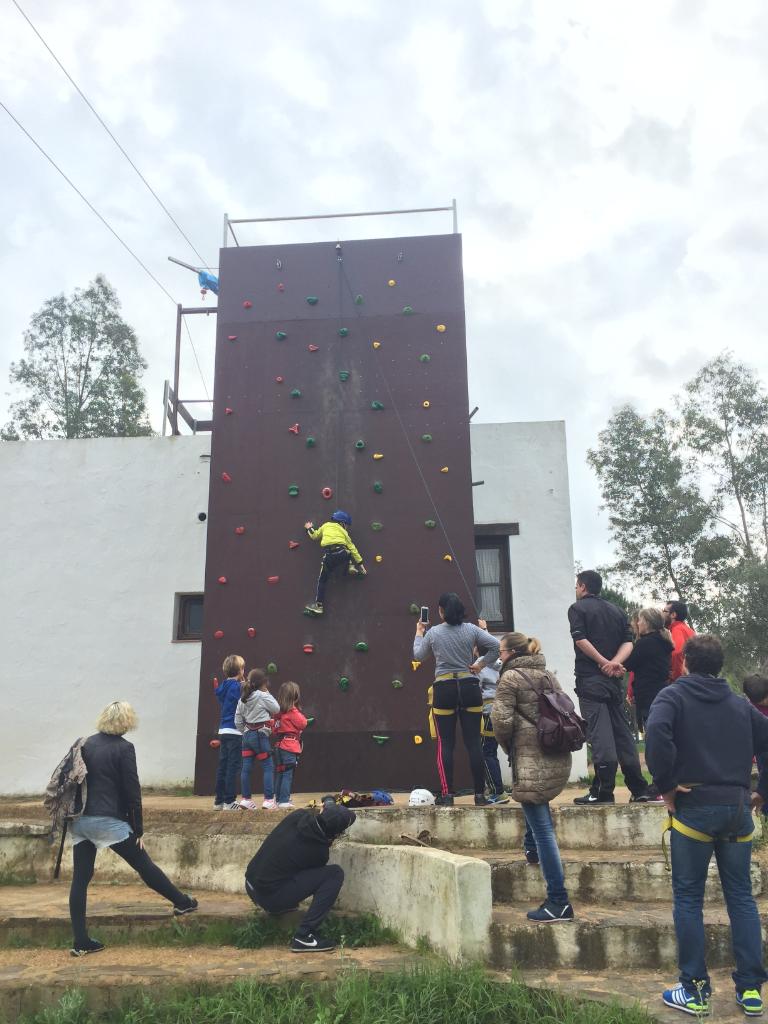 Un niño con ropa deportiva y arnés de seguridad escala un muro artificial mientras un grupo de personas observa y anima desde el suelo