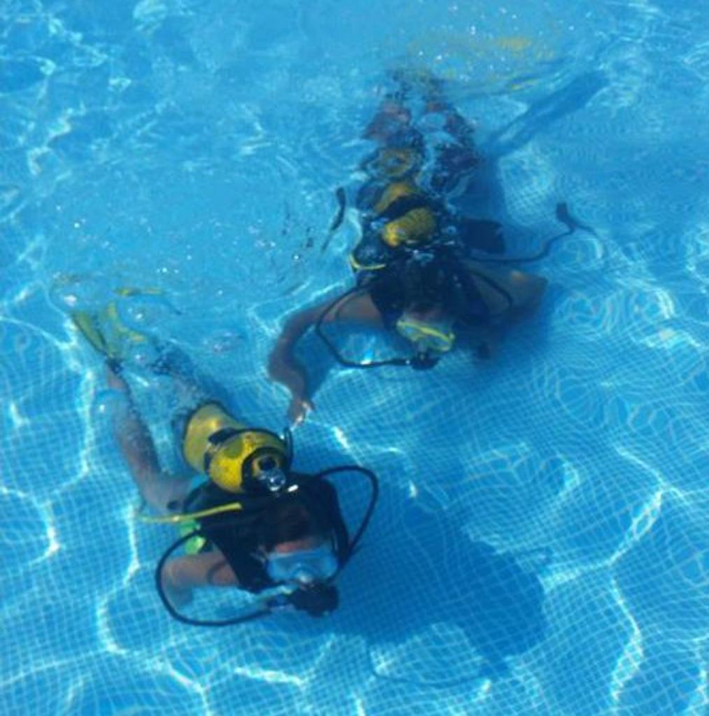 Dos personas con equipo de buceo practican en una piscina de agua cristalina, aprendiendo técnicas de inmersión en un entorno seguro