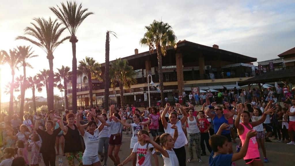 Gran grupo de personas de todas las edades participa en una animada sesión de baile al atardecer, rodeados de palmeras y un ambiente festivo en el paseo marítimo