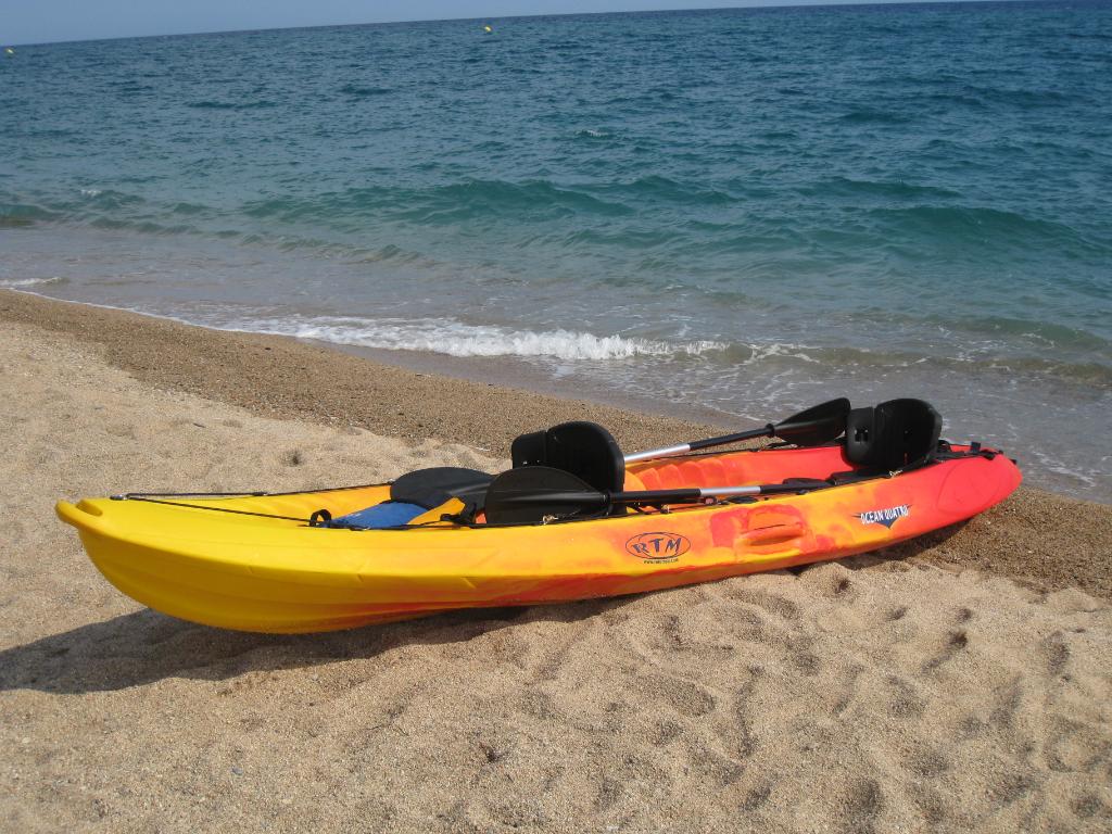 Kayak bicolor amarillo y rojo con remos negros descansando sobre la arena junto al mar, con pequeñas olas en la orilla y cielo despejado
